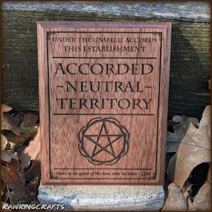 Puede incluir: Placa de madera rectangular marrón con texto grabado y símbolo de pentagrama. El texto dice "UNDER THE UNSEELIE ACCORDS THIS ESTABLISHMENT ACCORDED -NEUTRAL- TERRITORY" y "There is no spirit of the law, only its letter - OM".