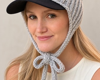 May include: A black baseball cap featuring a light gray crocheted earflap attachment that wraps around the sides and secures under the chin with a matching gray crocheted tie. The cap has a solid black brim and a black mesh crown.