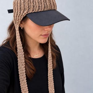 May include: A black baseball cap featuring a beige crocheted earflap attachment that wraps around the crown and extends into two long, dangling crocheted straps on either side of the face. The cap has a solid black brim and mesh back panels.