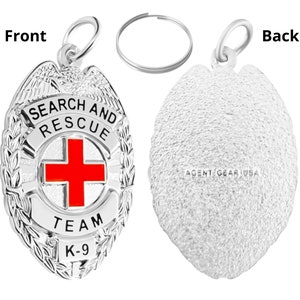 Op de afbeelding: Zilverkleurige metalen sleutelhanger met een zoek- en reddingsbadge. De badge heeft een rood kruis, de woorden "Search and Rescue", "Team" en "K-9", en een laurierkransontwerp. De achterkant van de sleutelhanger is effen met de woorden "Agent Gear USA" erop gegraveerd.