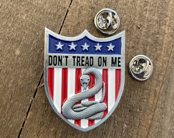 Pode incluir: Um broche de metal prateado em forma de escudo com um design de bandeira americana vermelha, branca e azul. O escudo apresenta uma cascavel enrolada com o texto "Don't Tread On Me" acima.