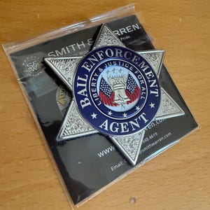 May include: A silver-colored, star-shaped badge with the words "BAIL ENFORCEMENT AGENT" in blue and white. The center features the Liberty Bell and American flags. The badge is on a black card with the "SMITH & WARREN" logo.