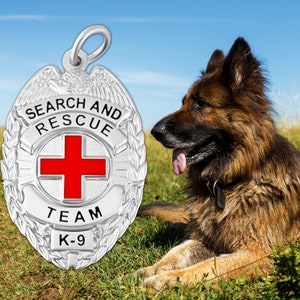 Op de afbeelding: Zilveren zoek- en reddingsbadge met een rood kruis, de woorden "Search and Rescue" en "Team K-9" en een laurierkransontwerp. Een Duitse herder zit in het gras.