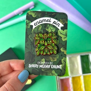 May include: Green enamel pin featuring a green man face with a red beard and brown eyes. The pin is on a card with a green and brown leafy background. The card reads "enamel pin" and "Designed by Stacey McEvoy Caunt".