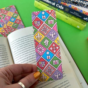May include: A colourful bookmark with a repeating pattern of medieval-style crests and symbols. The bookmark is pink, blue, yellow, green, and red.