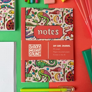 May include: A collection of notebooks and stationery on a green and red surface. The notebooks feature a colorful fantasy illustration with dragons and other creatures. One notebook has the word "notes" on the cover. The notebooks are labeled "Dot Grid Journal".