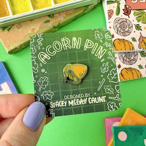 Peut inclure: Une épingle en forme de gland sur une carte verte avec le texte "ACORN PIN" et "DESIGNED BY STACEY MCEVOY CAUNT." L'épingle est jaune et verte. Le fond est vert avec d'autres articles de papeterie.