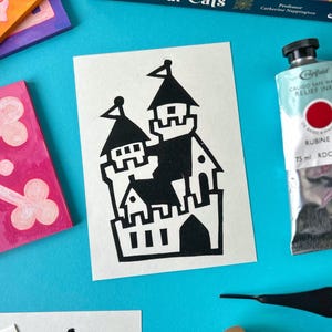 May include: A black and white illustration of a castle on a white card. The castle is a simple design with a tall tower and a few smaller towers. The illustration is surrounded by a white border.