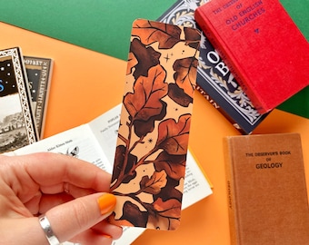 May include: A bookmark with a fall-themed design featuring brown and orange leaves with a white background. The leaves are stylized and have a vintage look. The bookmark is being held by a hand.