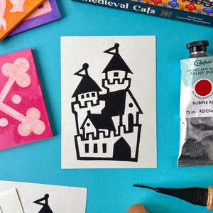 May include: Black and white linocut print of a castle with a flag on top. The castle is made up of multiple towers and has a gate with three windows. The print is on white paper.