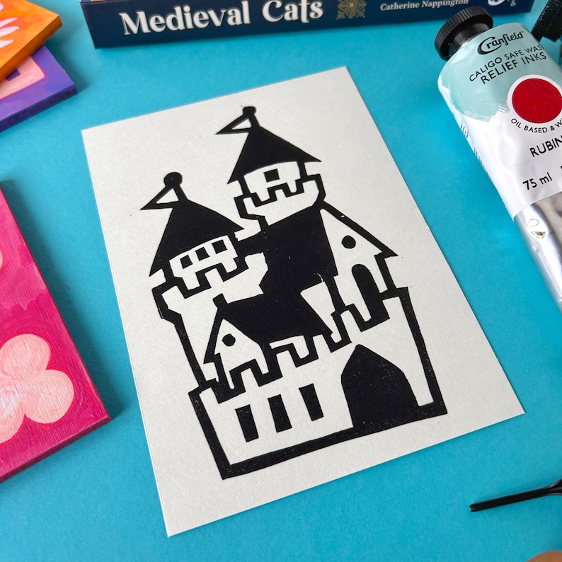 May include: Black and white linocut print of a medieval castle with a flag on top. The print is on a white card.