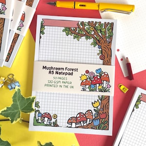 May include: A rectangular A5 notepad with a grid pattern and a decorative border featuring brown tree trunks, red and white mushrooms, and colorful flowers. A paper band displays the text Mushroom Forest A5 Notepad, 50 pages, 120 GSM paper, printed in the UK.