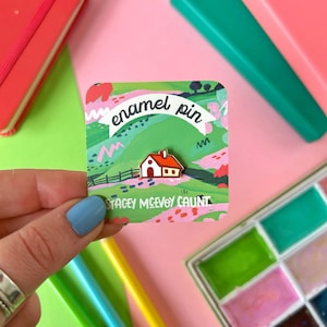 May include: A red enamel pin featuring a small white house with a red roof. The pin is on a card with a green and pink landscape design and the text "enamel pin" and "Stacey McEvoy Caust".