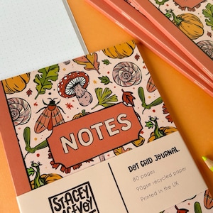 May include: A pink and orange dot grid journal with a fall-themed cover featuring mushrooms, pumpkins, and leaves. The cover also has the text "NOTES" in a large, bold font. The journal is 80 pages and made with 90gsm recycled paper. It is printed in the UK.