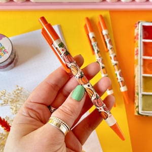 May include: Orange pens with a pumpkin pattern and white ink. The pens are on a yellow surface with a watercolor paint set in the background.