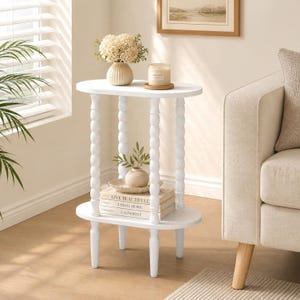 May include: A white oval side table with four spindle-style legs. The top surface holds a cream-colored vase with white flowers and a candle. The bottom shelf holds a small vase and a stack of three books titled Live Beautifully, This Is Home, and Calderone.