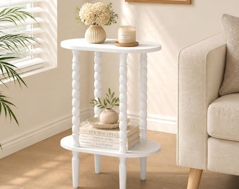 May include: A white oval side table with four spindle-style legs. The top surface holds a cream-colored vase with white flowers and a candle. The bottom shelf holds a small vase and a stack of three books titled Live Beautifully, This Is Home, and Calderone.