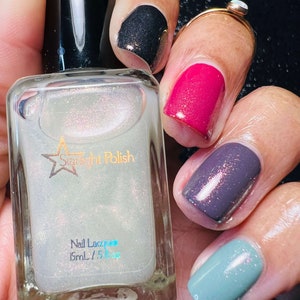 May include: A clear glass bottle of nail polish with a white label that says "Starlight Polish" and "Nail Lacquer 15mL / .5 fl oz". The bottle is being held by a hand with several fingernails painted in different colors, including black, pink, purple, and blue.