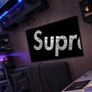Puede incluir: Una pieza de arte mural rectangular que presenta la palabra Supreme en letras grandes, negritas y blancas. Las letras están rellenas con un patrón de periódico en blanco y negro, sobre un fondo negro sólido dentro de un marco negro fino.