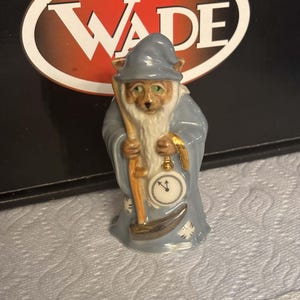 Pode incluir: Uma estatueta de cerâmica de uma raposa vestida de mago com uma túnica azul e chapéu pontudo, segurando um cajado de madeira e um relógio de bolso branco com ponteiros pretos. A raposa tem olhos verdes e uma longa barba branca.