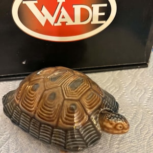 Könnte beinhalten: Eine kleine Schildkrötenfigur aus Keramik mit einem glänzenden braun-grauen Panzer, der ein detailliertes sechseckiges Muster aufweist. Die Schildkröte hat einen glatten, hellbraunen Kopf und steht vor einer schwarzen Schachtel mit einem weiß-roten ovalen Wade-Logo.