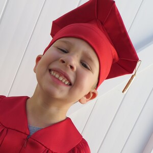 以下が含まれることがあります： 赤い卒業帽子とガウンを着て笑顔の子供。帽子には金のタッセルが付いています。ガウンには襟とジッパーが付いています。背景は白いスラットの壁です。子供はカメラを見ています。