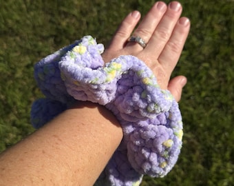 May include: A plush, oversized scrunchie made from soft, textured yarn in a light lavender purple color, featuring subtle flecks of yellow and green throughout the fabric, creating a thick, ruffled appearance around the wrist.