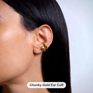 Peut inclure: Une boucle d'oreille en or massif avec une finition lisse et polie et un design ouvert et incurvé. Le bijou est porté sur le cartilage de l'oreille. Le texte "Chunky Gold Ear Cuff" est affiché sur une étiquette blanche en bas.