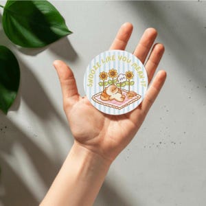 May include: A circular sticker featuring a light blue and white striped background with a cartoon cat sleeping on a pink checkered blanket. The illustration includes sunflowers and the text SNOOZE LIKE YOU MEAN IT in yellow letters.
