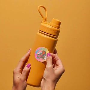 May include: A matte mustard yellow water bottle with a carrying loop on the cap. A circular sticker is applied to the center, featuring a pink roller skate, white daisies, and the text "Let's roll baby" in a light blue cursive font.