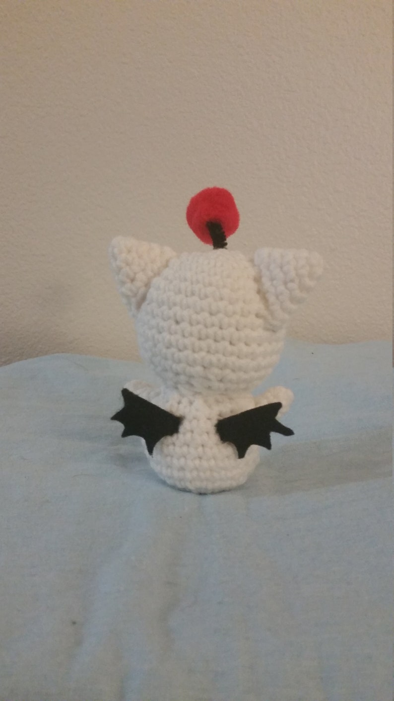 Puede incluir: Juguete de peluche blanco tejido a crochet con alas negras y un pomp&oacute;n rojo en la parte superior de la cabeza.
