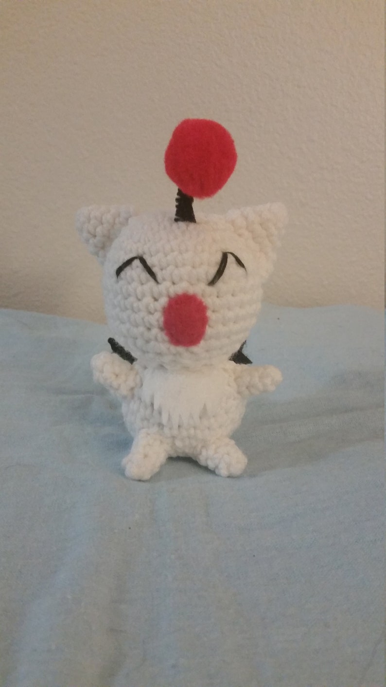 Puede incluir: Juguete de peluche blanco tejido a crochet con un pomp&oacute;n rojo en la parte superior de la cabeza. El juguete tiene ojos negros y una nariz roja.