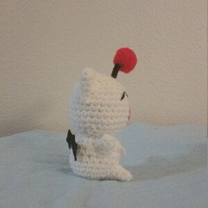 Puede incluir: Juguete de peluche blanco tejido a crochet con ojos negros y un pomp&oacute;n rojo en la parte superior de su cabeza. El juguete est&aacute; sentado sobre una superficie azul claro.