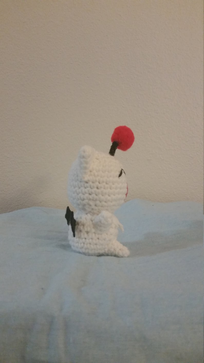 Puede incluir: Juguete de peluche blanco tejido a crochet con ojos negros y un pomp&oacute;n rojo en la parte superior de su cabeza. El juguete est&aacute; sentado sobre una superficie azul claro.