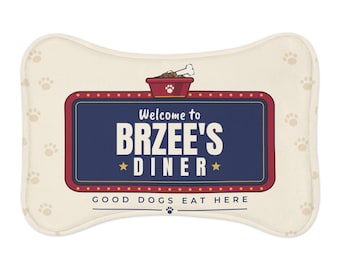 May include: A bone-shaped cream-coloured mat featuring a dark blue rectangular sign with a red border. The sign reads Welcome to BRZEE'S DINER with a dog bowl graphic above and the text GOOD DOGS EAT HERE below. The mat has light brown paw prints.