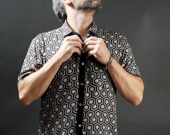 May include: A man wearing a black and white geometric patterned short-sleeved shirt and brown cargo shorts. He is buttoning the shirt with one hand.