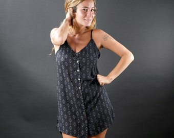 May include: A woman wearing a black and white patterned button-down dress with a black background and white geometric shapes. She is standing with her hands on her hips and is wearing black sandals.