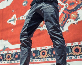 May include: A pair of gray pants with black detailing and a zipper on the side. The pants are worn with black ankle boots. The person is standing in front of a wall with a red, white, and black patterned design. The text '#R A' is visible on the wall.