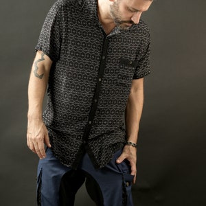 May include: A man wearing a black and white patterned short-sleeved shirt and blue denim shorts with black accents. The shorts have a large cargo pocket on the right side.