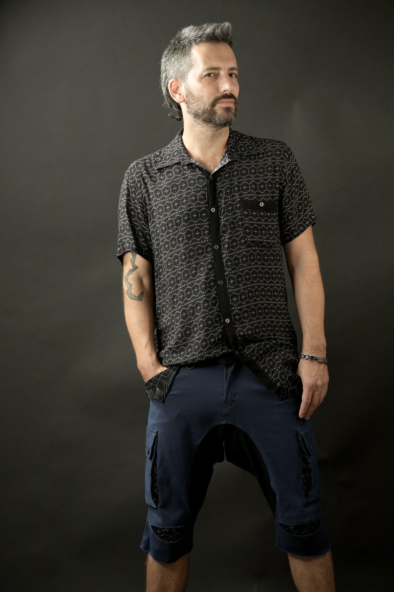 May include: A man wearing a black and white patterned short-sleeved shirt and dark blue shorts. He has a tattoo on his left arm.