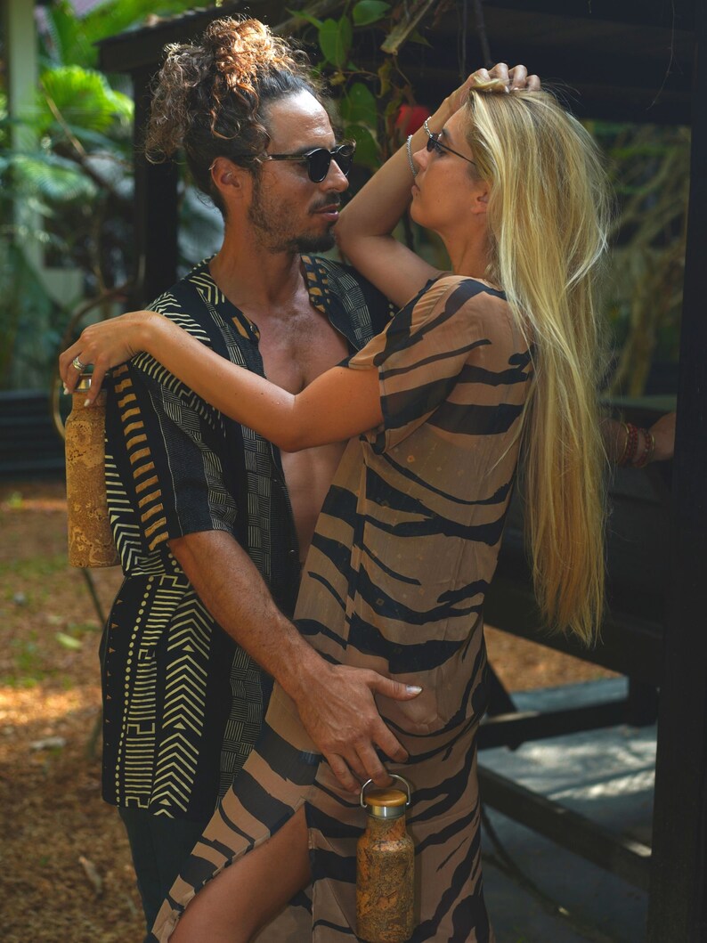 Op de afbeelding: Een stel staat dicht bij elkaar in een tropische omgeving. De vrouw draagt een bruin-zwarte gestreepte jurk en heeft lang blond haar. De man draagt een zwart-gouden geprint overhemd. Ze houden allebei bruine waterflessen vast.