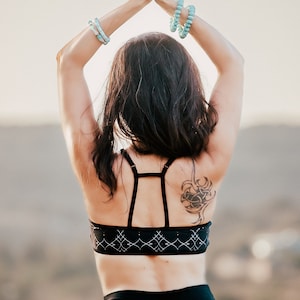 Op de afbeelding: Een vrouw draagt een zwarte sportbeha met een wit geometrisch patroon en een zwarte legging. Ze heeft een tatoeage op haar rechter schouderblad. Haar armen zijn boven haar hoofd geheven met haar handen in elkaar gevouwen.