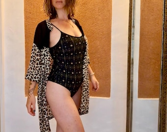 Op de afbeelding: Een zwart eendelig rompertje met een subtiel geometrisch patroon in goud en wit, gecombineerd met een open kimono-stijl overgooier. De overgooier heeft een effen zwart bovendeel en een luipaardprint op het onderste deel en de mouwen.