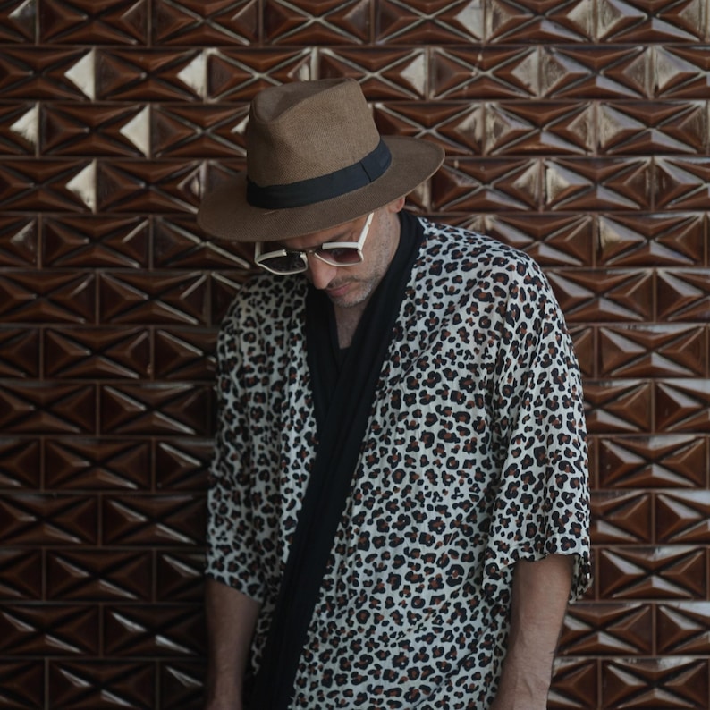 May include: A person wearing a brown fedora hat with a black band, white sunglasses, and a leopard print kimono-style jacket. A black scarf is draped around the neck. The background is a brown geometric tile wall.