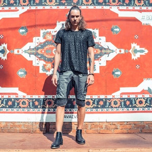 May include: A black short-sleeved t-shirt with a repeating geometric pattern, paired with grey cargo shorts featuring side pockets and black leather ankle boots. The outfit is worn by a person with long hair standing in front of a red wall.