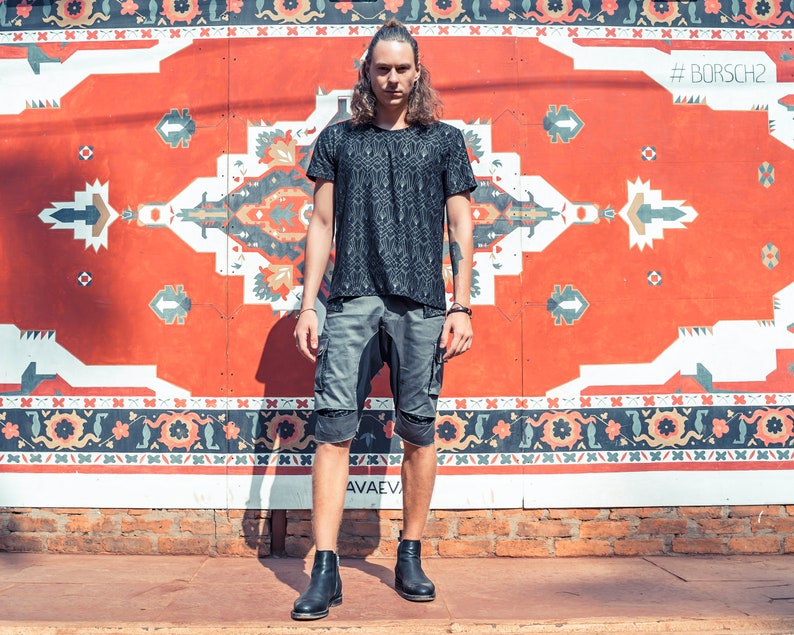 May include: A black short-sleeved t-shirt with a repeating geometric pattern, paired with grey cargo shorts featuring side pockets and black leather ankle boots. The outfit is worn by a person with long hair standing in front of a red wall.