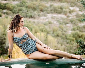Peut inclure: Une femme portant un maillot de bain à motifs bleus et bruns avec une découpe est assise au bord d'une piscine. La piscine est entourée de verdure.