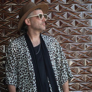 May include: A person wearing a brown fedora hat, white-framed sunglasses, and a leopard print kimono-style jacket. The person is also wearing a black shirt and a silver necklace. The background is a brown, textured wall.