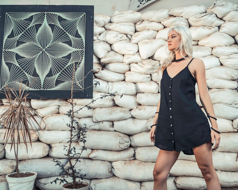 May include: A woman with long blonde hair wearing a black sleeveless dress with buttons down the front, standing in front of a wall of white sandbags.