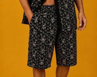 May include: A black short-sleeved button-down shirt and matching knee-length shorts featuring an all-over white graphic print of stylized eyes, geometric shapes, and arrows. The set is constructed from a dark fabric with a repeating pattern.